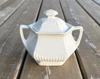 Può includere: Una zuccheriera in ceramica bianca con coperchio abbinato, caratterizzata da un corpo esagonale e due manici laterali. La base e il coperchio sono decorati con scanalature verticali e il coperchio è sormontato da un piccolo pomello a coste.