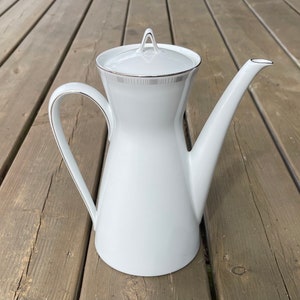 May include: A white porcelain coffee pot with a long spout and a curved handle. The pot has a lid and is trimmed with a silver-colored band around the top and handle. The pot is sitting on a wooden surface.