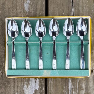 May include: A set of six silver spoons presented in a yellow and green box. Each spoon features a decorative floral pattern on the handle and a scalloped edge on the bowl. The spoons are neatly arranged within the box.