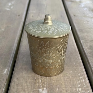 Peut inclure: Un récipient cylindrique en laiton avec un couvercle assorti, présentant un motif floral et végétal gravé sur toute la surface extérieure. Le couvercle est surmonté d'une petite poignée conique à gradins, avec une finition dorée métallique.
