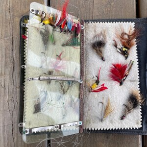 Puede incluir: Una caja de aparejos de pesca con mosca vintage de cuero negro abierta, llena de varias moscas de pesca coloridas. Las moscas presentan dise&ntilde;os de plumas rojas, amarillas y marrones. El interior presenta un forro de fieltro color crema y divisores de pl&aacute;stico transparente.