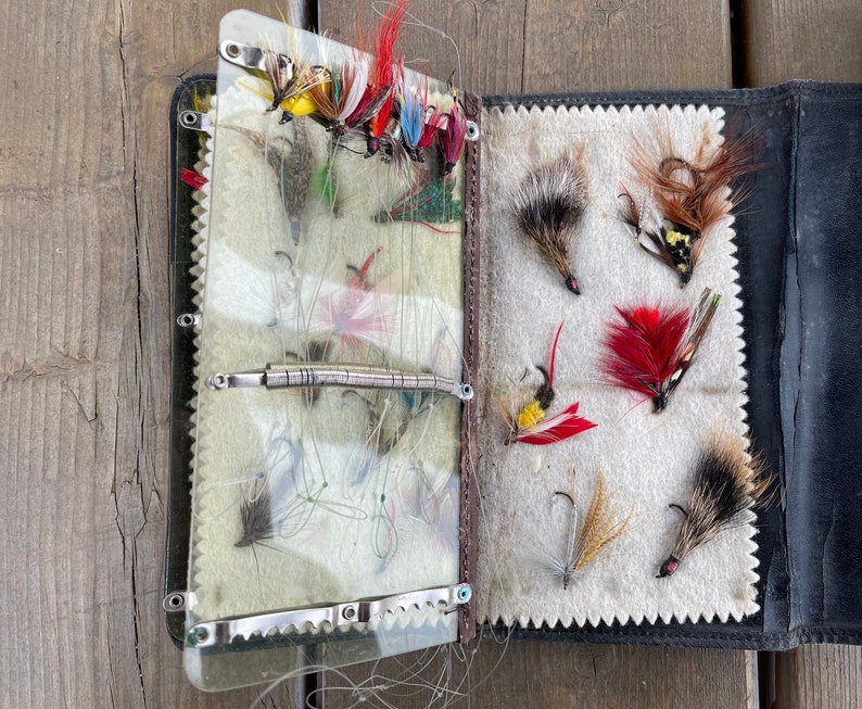 Puede incluir: Una caja de aparejos de pesca con mosca vintage de cuero negro abierta, llena de varias moscas de pesca coloridas. Las moscas presentan dise&ntilde;os de plumas rojas, amarillas y marrones. El interior presenta un forro de fieltro color crema y divisores de pl&aacute;stico transparente.