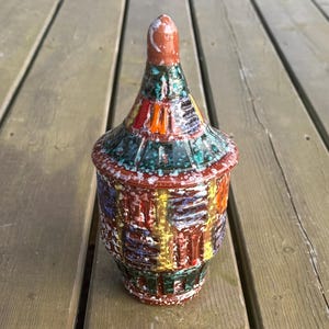 May include: A decorative ceramic jar with a lid. The jar has a patchwork design in red, green, orange, yellow, and white. The lid is a lighter red with a rounded top. The jar is on a wooden surface.