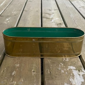 Peut inclure: Un cache-pot ovale en laiton avec un extérieur doré poli et un intérieur vert vif. Le récipient en métal présente une bande horizontale texturée qui longe les sections supérieure et inférieure de la paroi extérieure.