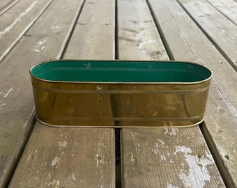 Può includere: Un portavaso ovale in ottone con esterno color oro lucido e interno verde brillante. Il contenitore in metallo presenta un dettaglio a fascia orizzontale testurizzata che corre lungo le sezioni superiore e inferiore della parete esterna.