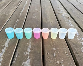 Può includere: Una fila di otto piccoli bicchieri in plastica traslucida con coperchi abbinati, disposti in linea. I bicchieri presentano una varietà di colori tra cui blu, azzurro, verde menta, rosa, pesca, giallo e due contenitori trasparenti.