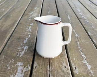 Op de afbeelding: Een witte keramische kan met een enkele donkerbruine horizontale lijn rond de bovenrand. De kan heeft een gebogen tuit en een handvat aan de zijkant met een gestructureerd, bamboe-achtig uiterlijk, wat zorgt voor een eenvoudig ontwerp.