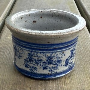 May include: A ceramic planter with a blue and white floral pattern. It has a wide, grey rim and a blue band. The exterior is speckled grey. The planter is on a wooden surface. The planter's diameter is approximately 15 cm.