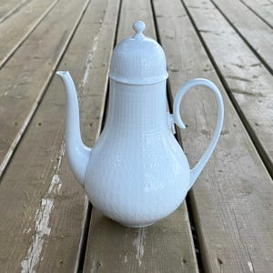 May include: A white ceramic coffee pot with a textured surface and a lid. The pot has a curved spout and a handle. The pot is sitting on a wooden surface.