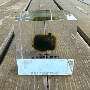 May include: A clear, transparent acrylic cube containing a dark, textured sample of oil sand from Fort McMurray, Alberta. The front face features black printed text that reads "OIL SAND" and "FORT MCMURRAY, ALBERTA" in a sans-serif font.