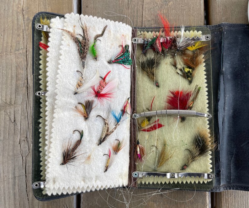 Puede incluir: Una caja de aparejos de pesca con mosca vintage abierta, que revela una selecci&oacute;n de moscas de pesca coloridas. La caja es negra con un interior verde. Las moscas est&aacute;n hechas de plumas y otros materiales, unidas a peque&ntilde;os anzuelos.