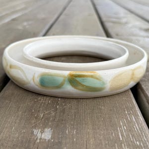 Peut inclure: Un bol en céramique blanc avec un design circulaire. Le bol a un anneau intérieur surélevé et est décoré de motifs en forme de feuilles vertes et beiges autour du bord. Le bol est posé sur une surface en bois.