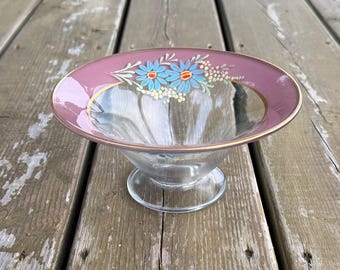 Op de afbeelding: Een glazen schaal op voet met een brede, mauvekleurige rand versierd met handgeschilderde lichtblauwe madeliefjes, oranje harten en kleine witte bloemaccenten. De schaal is voorzien van een goudkleurige rand langs de binnen- en buitenkant.