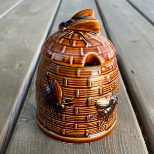 Op de afbeelding: Een keramische honingpot in de vorm van een bijenkorf met een bij op de deksel. De pot is bruin en heeft een getextureerd oppervlak.