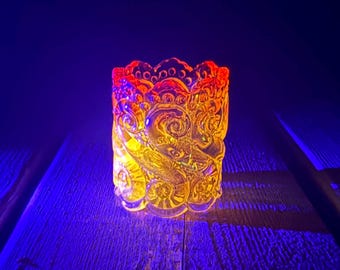 Op de afbeelding: Een votiefkaarshouder van helder glas met een verhoogd, wervelend reliëfpatroon van schelpen en op de oceaan geïnspireerde motieven. Het glas gloeit van binnenuit met een warm oranje licht, contrasterend met een donkere, blauwe omgeving.