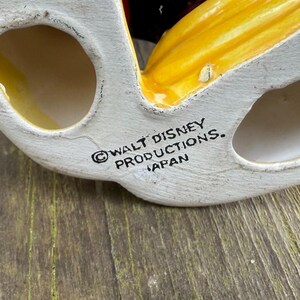 May include: A vintage ceramic figurine base, with a white base and yellow and red details. The text "WALT DISNEY PRODUCTIONS. JAPAN" is printed on the base. Likely a collectible item.