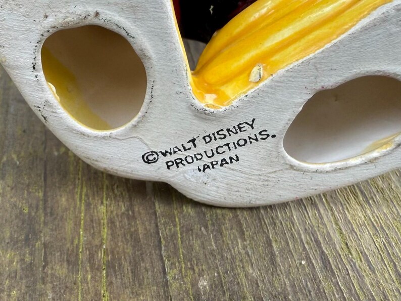 May include: A vintage ceramic figurine base, with a white base and yellow and red details. The text "WALT DISNEY PRODUCTIONS. JAPAN" is printed on the base. Likely a collectible item.