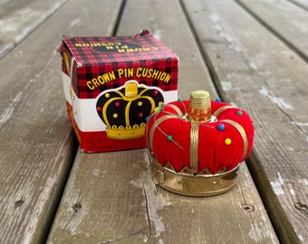 Può includere: Un puntaspilli in velluto rosso a forma di corona con base in metallo dorato e un ditale sulla parte superiore, accompagnato dalla scatola originale con l'etichetta CROWN PIN CUSHION e un motivo a quadri rossi e neri.