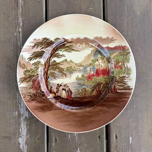 May include: Round decorative plate with a scenic landscape design. The plate features a brown border and a central image of a castle, trees, and figures near a body of water. The color palette includes brown, beige, red, and green.