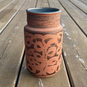 May include: A handcrafted ceramic vase with a reddish-brown exterior. The vase has a dark brown, repeating leaf pattern and a textured band near the top. The rim is a contrasting blue-grey colour. This decorative vase is suitable for home decor.