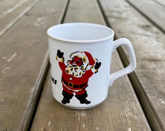Op de afbeelding: Een witte keramische mok met een grafische illustratie van de Kerstman in een rood pak met een zwarte riem en zwarte laarzen. De figuur heeft beide armen omhoog en de tekst Merry Christmas is gedeeltelijk zichtbaar aan de zijkant van de mok.