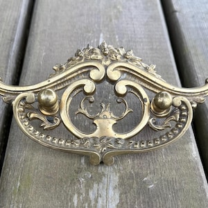 May include: A gold-toned brass drawer pull featuring an ornate, symmetrical design with scrolling curves, floral motifs at the top and bottom, and two small, rounded knobs positioned on either side of the central openwork frame.