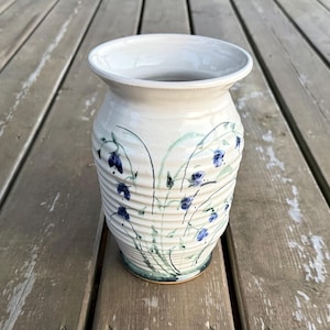 May include: A cream-coloured ceramic vase with a ribbed, horizontal texture. The exterior features a hand-painted botanical design with delicate green stems and small, scattered blue flowers. The vase has a wide, flared rim and a tapered base.