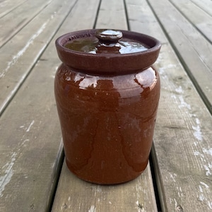 Peut inclure: Un pot en céramique marron de forme cylindrique avec une finition brillante et un couvercle assorti. Le récipient présente un large rebord et un petit bouton arrondi au centre du couvercle. La surface de la poterie est lisse et brillante.