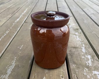 Op de afbeelding: Een cilindervormige bruine keramische pot met een glanzende afwerking en een bijpassend deksel. De houder heeft een brede rand en een kleine, ronde knop in het midden van het deksel. Het oppervlak van het aardewerk is glad en reflecterend.