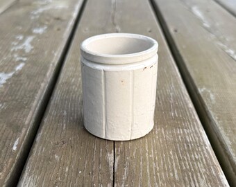 Op de afbeelding: Een cilindrische, gebroken witte keramische pot met verticale ribbels langs de buitenwanden en een gladde, open bovenrand. Het vat heeft een stevige basis en een uniforme, mat getextureerde afwerking met subtiele tekenen van ouderdom.