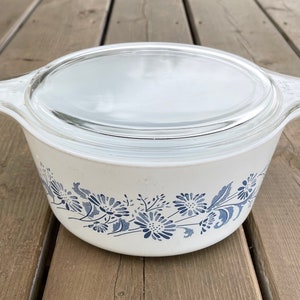 May include: A white casserole dish with a clear glass lid. The dish features a blue floral design around the exterior. The dish has two handles and is sitting on a wooden surface.
