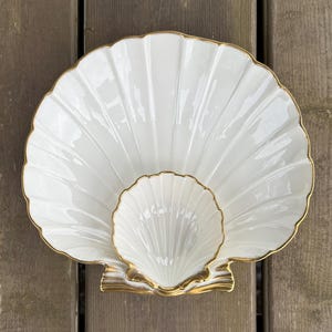 May include: A white, shell-shaped serving dish with a smaller dish nested inside. The edges are trimmed with gold. The dishes are made of porcelain and are set on a wooden surface.