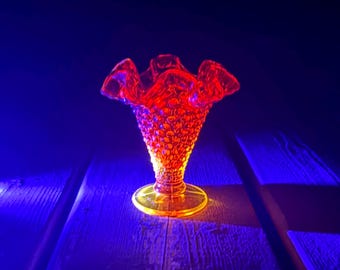 Op de afbeelding: Een feloranje glazen vaas met een hobnail-textuur en een gegolfde, geschulpte rand. De vaas wordt verlicht door een felle lichtbron, waardoor deze intens oplicht tegen een donkere, diepblauwe achtergrond.