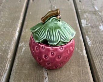 Può includere: Un barattolo in ceramica con coperchio a forma di fragola. La base è di un rosa scuro lucido con texture circolari incavate. Il coperchio presenta un design a foglie verdi lucide con un gambo marrone come manico sulla parte superiore.