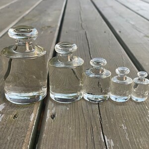 Peut inclure: Ensemble de cinq poids en verre transparent de tailles variées, disposés en ligne. Chaque poids a un corps arrondi et un petit dessus plat. Les poids sont numérotés, indiquant probablement leur poids en grammes.