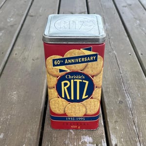 May include: A rectangular red metal Christie's RITZ cracker tin with a silver lid embossed with the word RITZ. The front features a 60th Anniversary graphic, the text 1935-1995, and a net weight of 450 g. The design includes images of round crackers.