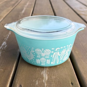 May include: A vintage turquoise Pyrex casserole dish with a clear glass lid. The dish features a white graphic design of a rooster, baskets, and people. The dish is sitting on a wooden surface.