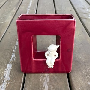 May include: A square, deep red ceramic vase with a glossy finish and a large central cutout. A small, white, matte-finished figurine of a seated elf with a pointed hat is attached to the bottom edge of the cutout, appearing to sit on the frame.