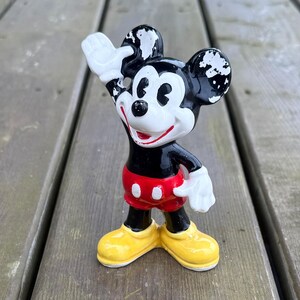 May include: A vintage Mickey Mouse figurine, with a black body, red shorts, yellow shoes, and white gloves. The figure is waving with one hand raised. The figurine is standing on a wooden surface.