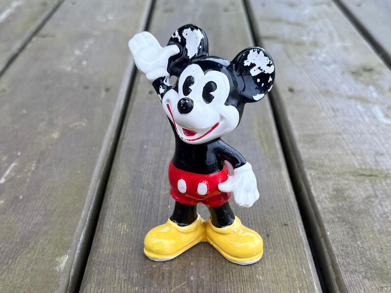May include: A vintage Mickey Mouse figurine, with a black body, red shorts, yellow shoes, and white gloves. The figure is waving with one hand raised. The figurine is standing on a wooden surface.