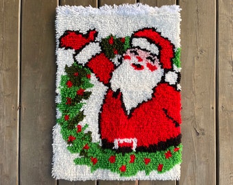 Op de afbeelding: Een rechthoekige, handgemaakte kerstwandhanger met de kerstman. De afbeelding is een close-up van een kerstmanontwerp met een rood pak, witte baard en een groene krans. De achtergrond is wit met een getextureerd, getuft uiterlijk.