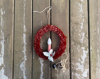 Può includere: Una ghirlanda circolare realizzata in materiale plastico rosso stropicciato con una candela elettrica centrale dotata di lampadina bianca e base rosa. Un accento a forma di foglia di agrifoglio argentata con una piccola bacca rossa si trova in basso.