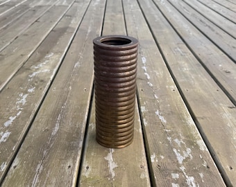 Può includere: Un vaso cilindrico con una finitura metallica marrone scuro e una prominente trama a coste, simile a una bobina, che corre verticalmente lungo l'intera superficie esterna. L'oggetto presenta una parte superiore circolare aperta e una base piatta.