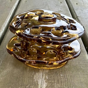 May include: Amber-colored glass candle holder with a unique, organic shape. The two-piece design features a base and a top section with multiple circular indentations. The glass has a glossy finish, reflecting light and creating a warm, inviting aesthetic.