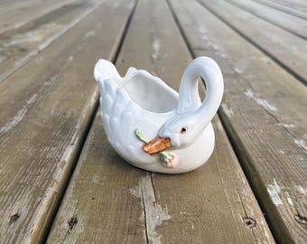 Può includere: Un vaso o piatto in ceramica bianca a forma di cigno con finitura lucida. Il cigno presenta un collo ricurvo, dettagli di piume modellate sul corpo e un becco arancione che tiene un piccolo fiore rosa con una foglia verde.