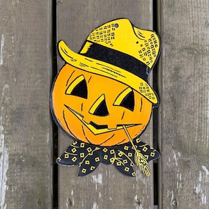 May include: A flat, decorative cutout of a jack-o'-lantern wearing a yellow hat with a black band. The pumpkin is orange with black triangular eyes, a nose, and a jagged mouth. A black neckerchief with a yellow diamond pattern is at the base.