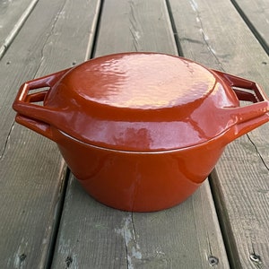 May include: A reddish-brown enameled cast iron Dutch oven with a matching lid. The pot has two handles and a silver rim. The lid has a textured surface. The cookware is designed for stovetop or oven use.