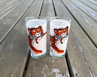 Può includere: Due bicchieri in vetro trasparente con un'illustrazione grafica di una tigre arancione e nera. La tigre è raffigurata con un muso bianco, strisce nere e un'espressione giocosa. I bicchieri hanno una forma cilindrica con bordo e base lisci.