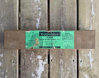 Op de afbeelding: Een rechthoekige bruine doos met 50 Gypsona gipsverbandplaten. Het groene etiket bevat de tekst Gypsona Slabs, inhoud 50 platen, 7,6 cm bij 76,2 cm, en gebruiksinstructies. De verpakking is gemaakt in Engeland door T.J. Smith & Nephew Ltd.