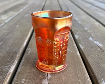Può includere: Un bicchiere in vetro iridescente color arancio calendula con un motivo stampato di farfalle e grappoli floreali. Il vetro presenta un bordo liscio svasato e una base circolare, con una finitura metallica riflettente che cattura la luce.